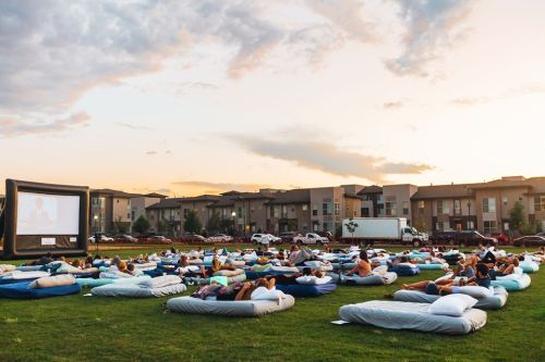 Outdoor movie night with people lounging on large cushions, watching a film on a big screen at sunset.