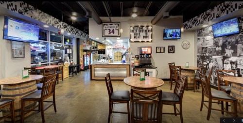 Interior of a casual restaurant with wooden tables, bar stools, and TVs displaying sports. Welcoming atmosphere.
