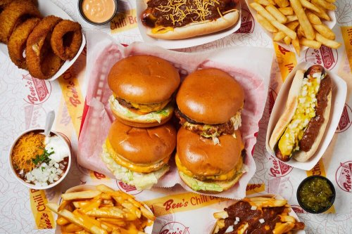 A spread of burgers, hot dogs, fries, onion rings, and toppings on a table with a red and white patterned background.