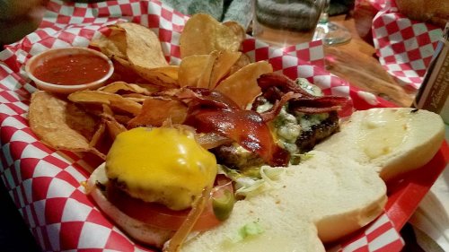 A plate with two burgers, one topped with cheese and bacon, served with chips and a side of salsa.