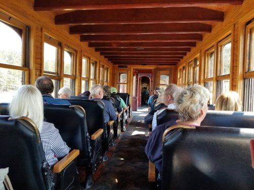 Interior of a vintage train car with passengers seated, wooden paneling, and large windows letting in natural light.