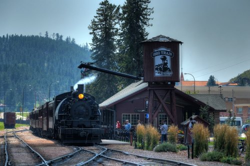 A vintage steam train at a station, with a water tower and trees in the background, under a cloudy sky.