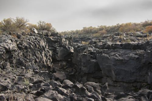 A rugged landscape of dark, jagged volcanic rock surrounded by sparse vegetation under a cloudy sky.