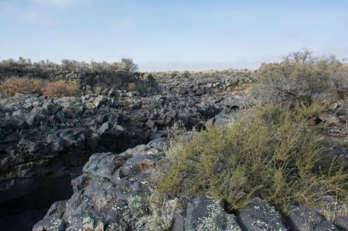A rocky landscape with dark volcanic formations and sparse vegetation under a clear sky.
