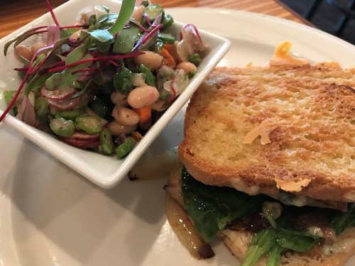 A toasted sandwich with greens and a side of colorful bean salad in a white bowl.