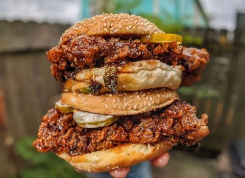 A towering sandwich with three layers of crispy fried chicken, pickles, and sesame seed buns, held in a hand.
