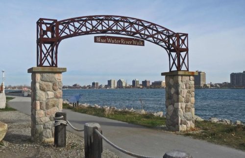 Archway sign for "Blue Water River Walk" with a scenic view of the river and city skyline in the background.