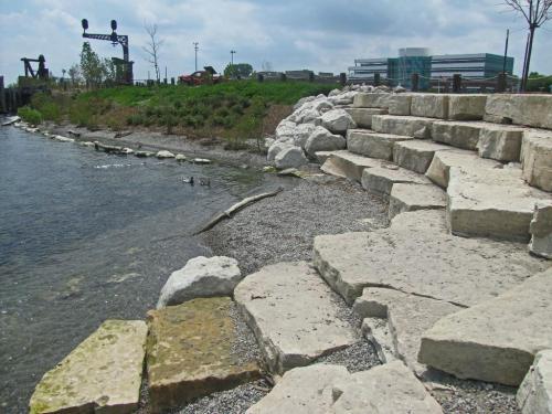 Rocky shoreline with steps leading to water, greenery in the background, and a modern building in the distance.