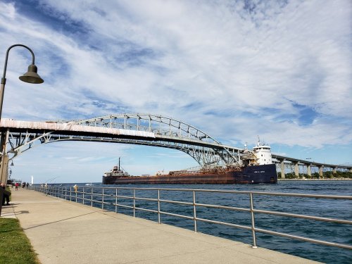 A large ship passes under a steel bridge, with a walkway and lamp post along the water's edge under a blue sky.