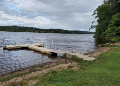 A calm lake with a small dock extending into the water, surrounded by trees and a cloudy sky.