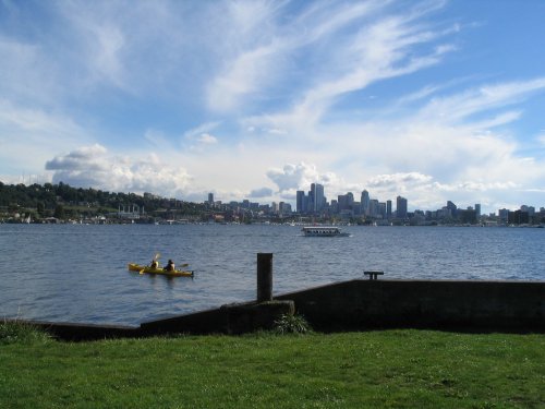 A serene view of a city skyline across a lake, with two kayakers in the foreground and fluffy clouds in the sky.