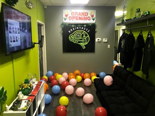 A vibrant room decorated for a grand opening, featuring a sign, balloons on the floor, and a black couch.