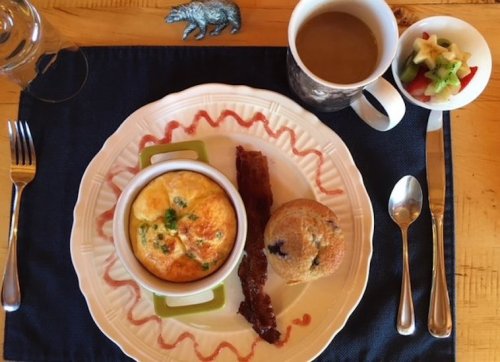 A breakfast plate with an omelet, bacon, a muffin, and a fruit cup, accompanied by a cup of coffee on a blue placemat.