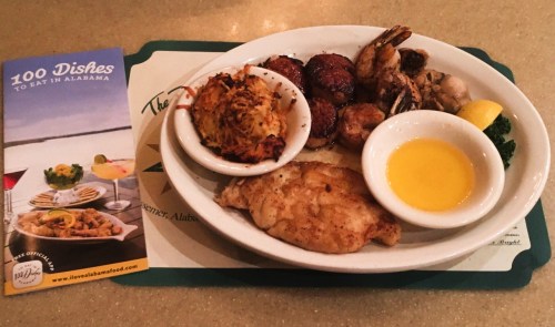 A plate of seafood including fish, shrimp, and crab cakes, accompanied by a lemon wedge and a brochure about Alabama dishes.