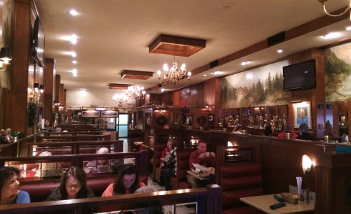 A cozy restaurant interior with wooden decor, chandeliers, and patrons enjoying meals at booths and tables.