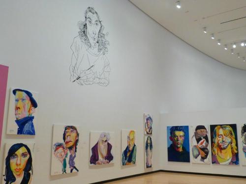 A gallery wall featuring colorful portraits and a large sketch of a person, showcasing diverse artistic styles.