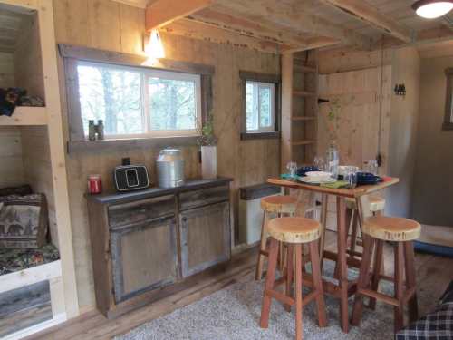 Cozy interior of a cabin with wooden decor, a dining table, and shelves, featuring natural light from windows.