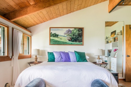Cozy bedroom with a wooden ceiling, a bed with colorful pillows, and a landscape painting above it.