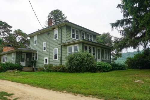 A green two-story house with a porch, surrounded by trees and grass, along a dirt road.