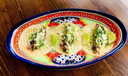 Three tacos on a colorful plate, topped with onions, cilantro, and lime wedges, set on a wooden table.