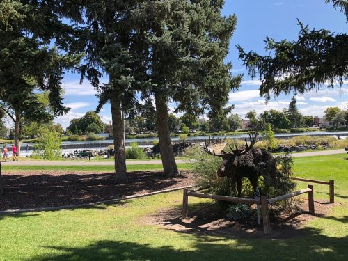 A park scene featuring a moose statue, trees, and a lake with people walking in the background under a blue sky.