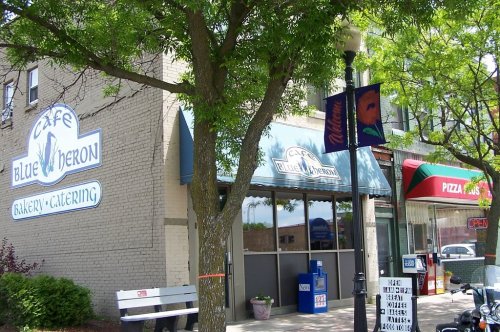Exterior of Café Blue Heron, featuring a bakery and catering sign, with trees and nearby shops in a vibrant street setting.