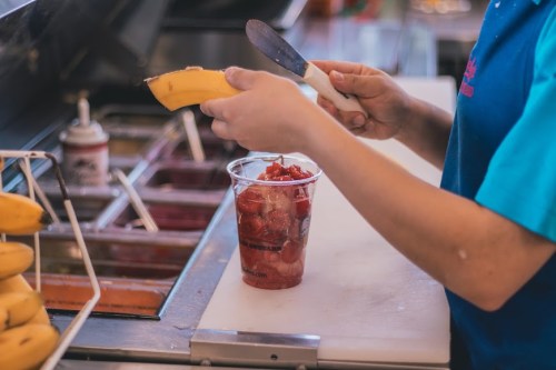 A person prepares a banana while holding a cup of strawberries at a food counter.