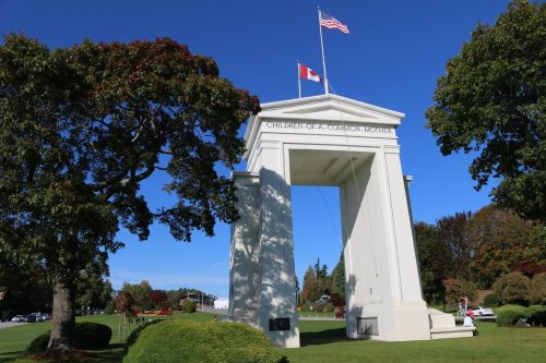 A white archway with flags of the U.S. and Canada, surrounded by green trees and blue sky, labeled "Children of a Common Mother."
