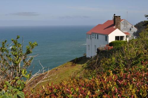 A white house with a red roof perched on a cliff overlooking the ocean, surrounded by greenery and coastal scenery.