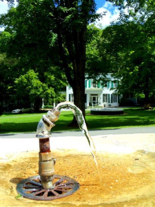 A water fountain spouts water in a park, with a large white building and green trees in the background.