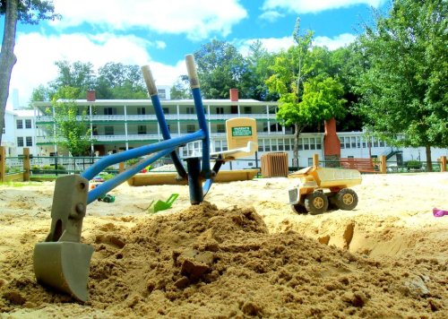 A sandbox with shovels and a toy truck, surrounded by trees and a building in the background under a blue sky.
