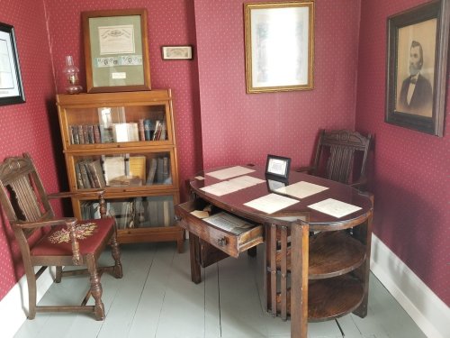 A vintage office with a wooden desk, bookshelves, and red wallpaper, featuring framed pictures and documents.