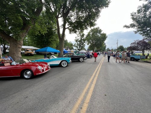 A street lined with classic cars and people walking, under trees with tents in the background on a cloudy day.