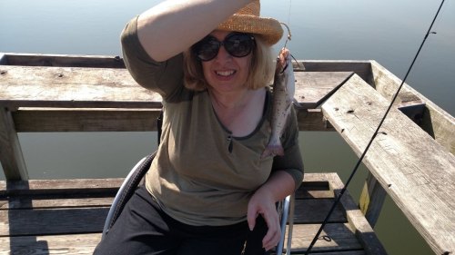 A smiling woman wearing a straw hat and sunglasses holds a fish while sitting on a wooden dock by the water.