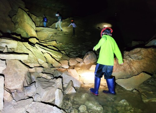 A group of people exploring a dark cave, walking over rocky terrain with headlamps and waterproof boots.