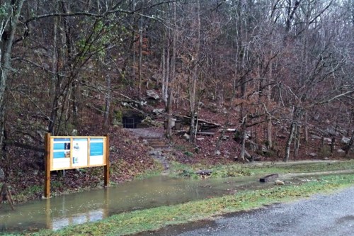 A wet trail leads to a rocky area, with a signboard in the foreground and bare trees in a rainy landscape.