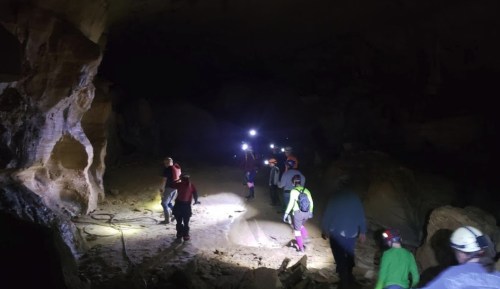 A group of people exploring a dark cave, equipped with helmets and headlamps, walking along rocky terrain.