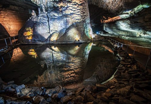 A cavernous space with illuminated rock formations and a still water pool reflecting the surroundings.