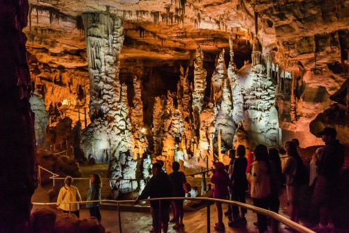 Visitors explore a stunning cave with towering stalagmites and stalactites, illuminated by warm lighting.