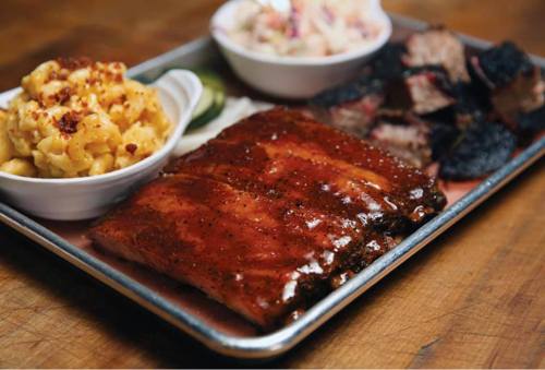 A platter featuring glazed ribs, smoked brisket, macaroni and cheese, and coleslaw, garnished with pickles.