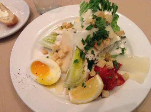 A fresh salad with romaine lettuce, croutons, a soft-boiled egg, tomatoes, lemon, and dressing on a white plate.