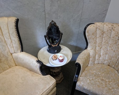 Two elegant chairs beside a small table with a cupcake and a cookie on a plate, set against a marble backdrop.