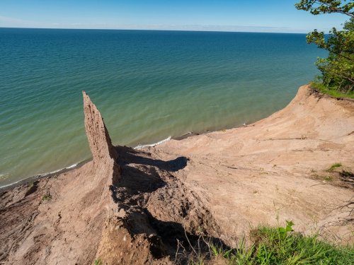 A steep, sandy cliff juts out over a calm lake, with clear blue skies above and gentle waves below.
