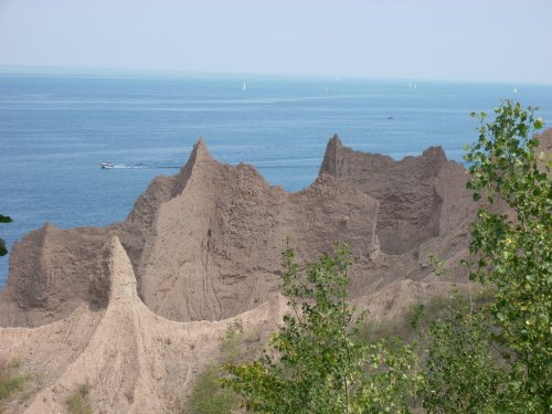 A rocky shoreline with jagged cliffs overlooking a calm blue sea, dotted with small boats in the distance.