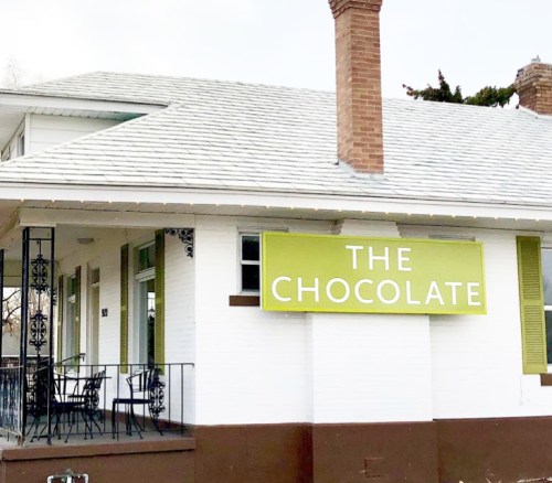 A white building with a green sign reading "THE CHOCOLATE" and a porch with chairs.