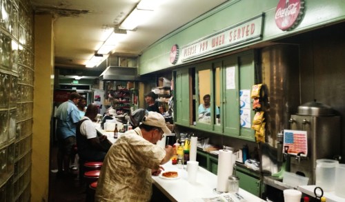 A cozy diner interior with patrons at the counter, vintage decor, and a menu sign overhead.