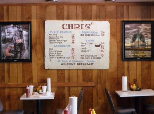 Interior of a diner featuring a menu board, wooden walls, and framed photos, with condiments on tables.