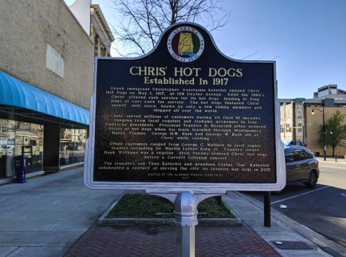 Sign for Chris' Hot Dogs, established in 1917, detailing its history and founders in a downtown setting.