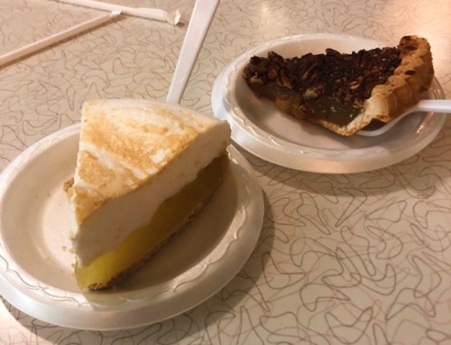 A slice of lemon meringue pie and a slice of pecan pie on paper plates, with plastic spoons beside them.