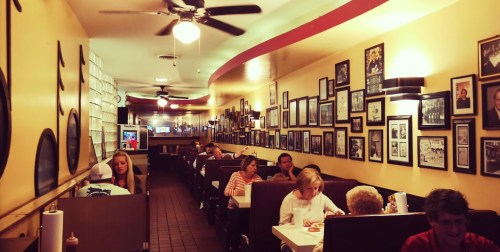 A cozy restaurant interior with diners at tables, walls adorned with framed photos, and ceiling fans overhead.